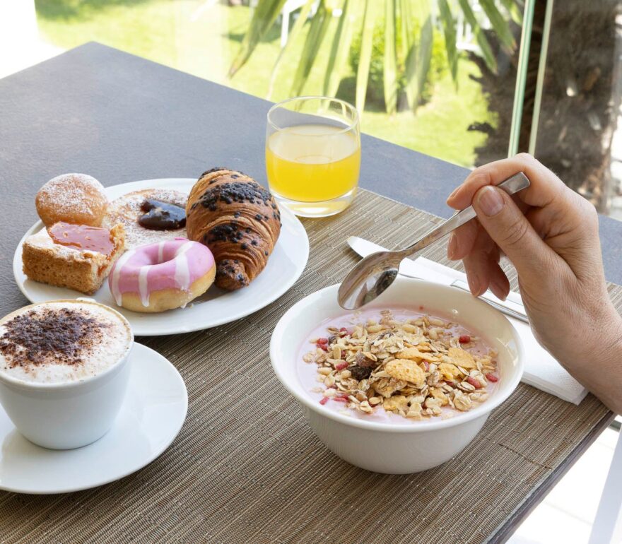 Una donna in accappatoio sta facendo colazione all'aperto su un tavolo marrone; ci sono vari dolci, una tazza di cappuccino e un bicchiere di succo d'arancia. Sullo sfondo si intravede un giardino con palme.